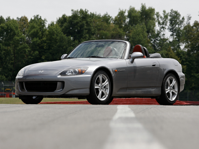 A 2009 Silver Honda S2000 convertible parked on the racetrack for a stationary picture with forest in the background.